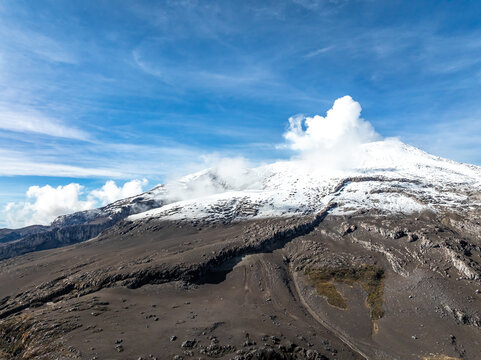 Aerial view of the snow-capped Nevado del Ruiz volcano with volcanic ash slopes, rocky terrain, and a plume of smoke under a bright blue sky in Villamaria, Caldas, Colombia.