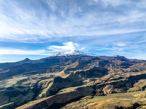 Aerial view of the snow-capped Nevado del Ruiz volcano with a plume of volcanic gas rising into a blue sky above the rugged Andean landscape of Villamaria, Caldas, Colombia.