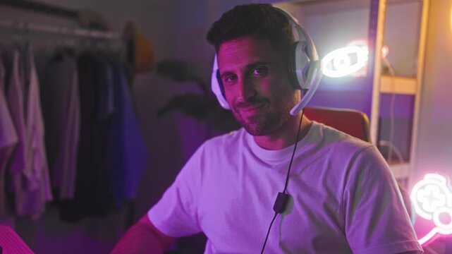 Man wearing white headset smiles at camera while seated in a studio gaming setup with neon signs, controller art and visible clothing rack; relaxed focus.