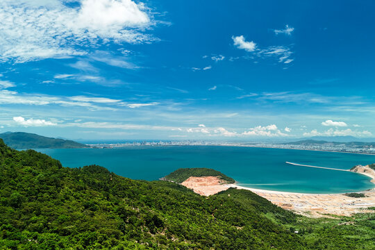 Coastal panorama from forested mountains overlooking turquoise sea, distant shoreline and waterfront development near Da Nang, Vietnam, under blue skies.