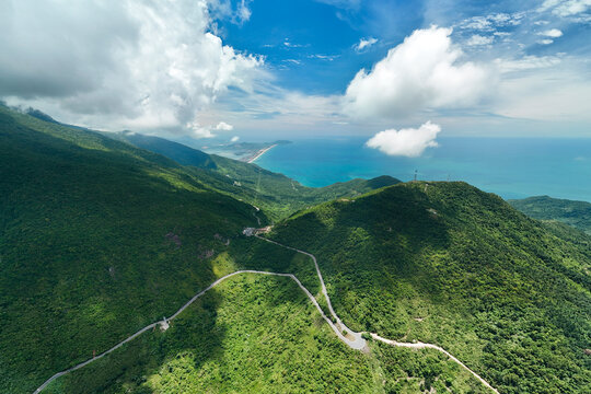 Aerial view across forested mountain ridges toward the turquoise coast, with a narrow winding road and scattered clouds under a vivid blue sky in Vietnam.
