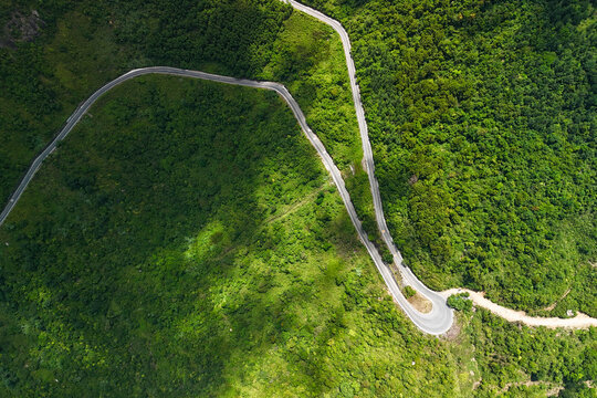 High-angle view of a serpentine mountain road cutting through dense tropical forest, with sharp bends and sunlit vegetation near Hai Van Pass, Vietnam.