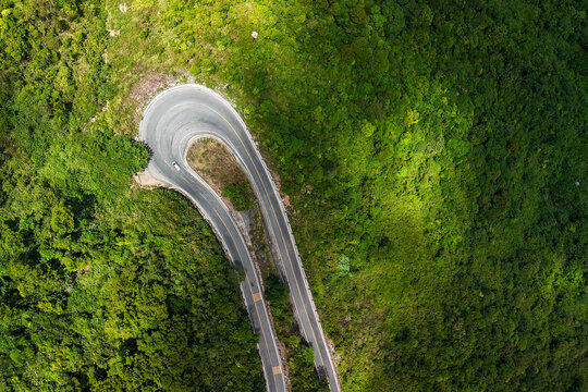 Aerial view of a tight hairpin bend on a mountain road surrounded by dense tropical forest, with a small vehicle navigating the curve at Hai Van Pass, Vietnam.