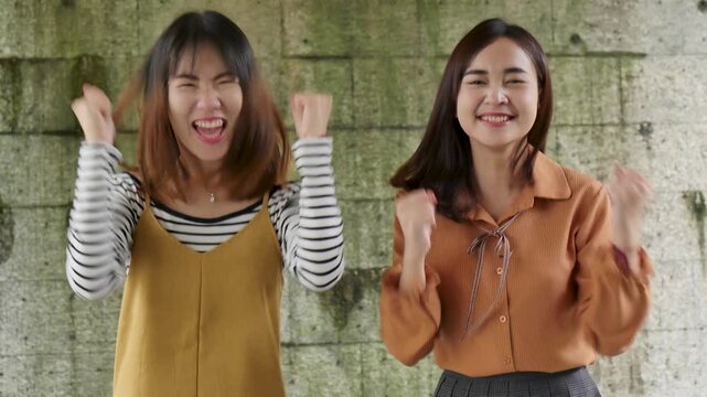 Two 20s Southeast Asian women laughing joyfully and doing fist pump together outdoors, celebrating friendship and success