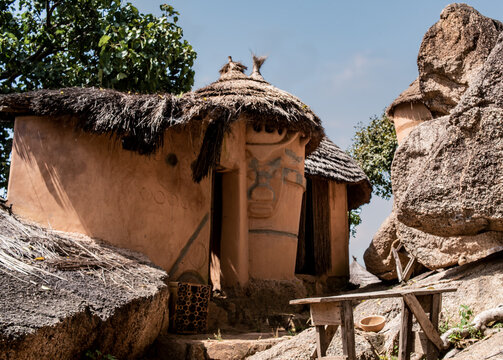 View of a traditional mud hut with a thatched roof and intricate wall carvings at the Museum of Traditional Architecture, nestled among large granite boulders in Jos, Plateau, Nigeria.