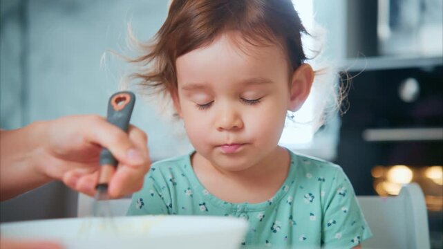 Soft light glows in kitchen. Child watches mixture being poured with quiet curiosity. Parent's hand guides spoon gently. Child's cheeks show mild concentration