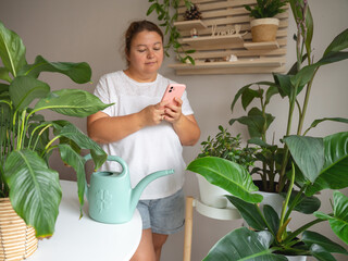 A woman stands in a bright room surrounded by lush green houseplants, using her smartphone while taking a break from gardening. This image represents plant care, digital lifestyle, and home hobbies. © JULIA