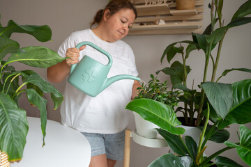 A middle-aged woman waters lush green houseplants with a light green watering can in a bright home interior, representing domestic wellness, urban gardening, and a sustainable lifestyle. © JULIA