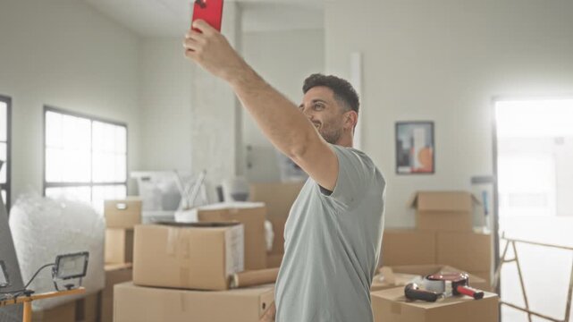 Man holding smartphone high taking a selfie amid cardboard moving boxes in building; optimism new beginning.