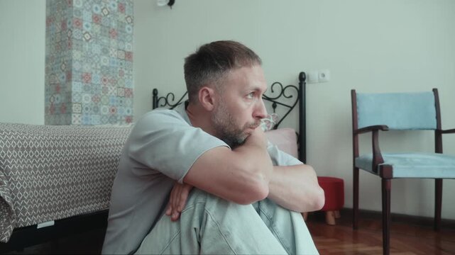 Man experiencing loneliness hugs knees on floor beside bed, isolated at home while facing sadness, depression, emptiness and growing emotional disconnection.