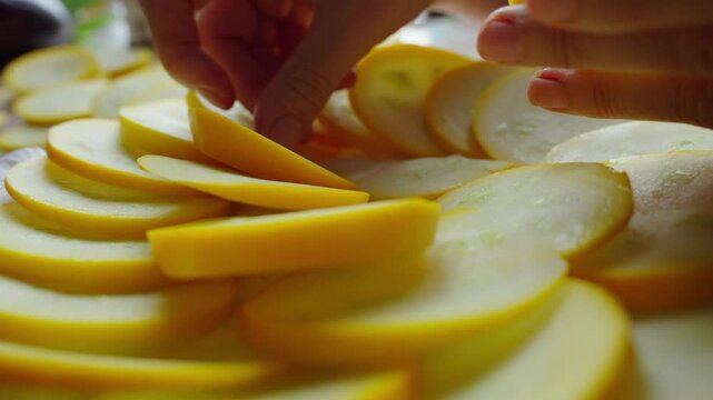 Hands carefully arrange yellow squash slices in a circular pattern. Fresh, crisp slices lie flat on a clean white surface. Natural light highlights the bright, even color of each round slice