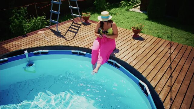 Woman relaxes on wooden deck beside pool. She wears bright pink pants and a straw hat. She sips drink while sitting near water. Her feet dip into clear blue pool water