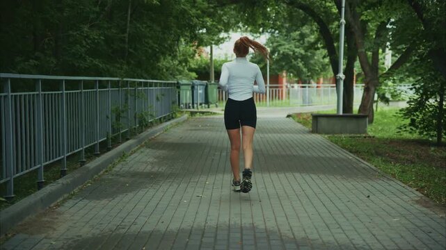 Young woman jogs on paved path through park. Trees line walkway behind her. She moves with steady rhythm. Path runs beside metal fence. Natural daylight illuminates scene. Ideal for fitness, wellness