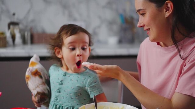 Mother feeds child with spoon in kitchen. Child holds plush toy while eating. Warm, caring moment between parent and child. Ideal for parenting or family content
