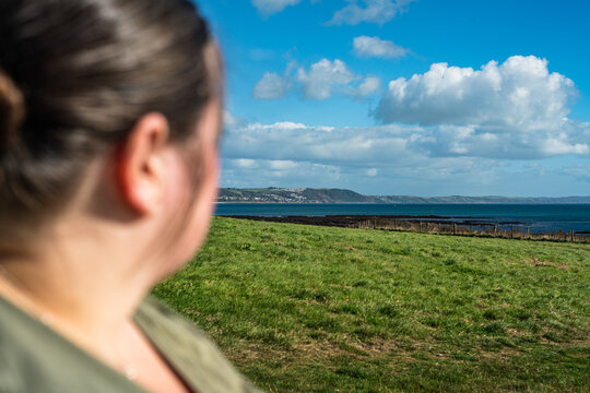 View of a woman in the foreground looking out over a green grassy field towards the sea and coastline under a blue sky with clouds in Looe, England, United Kingdom.
