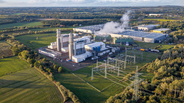 Aerial view of a combined-cycle gas turbine power plant and substation, Hamm North Rhine-Westphalia, surrounded by green fields and industrial warehouses, industrial zone.