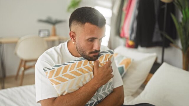 Man hugging patterned pillow on bed in a bedroom, hand clutching cushion, head bowed and pensive gaze, soft morning light falling on face; melancholy reflection.