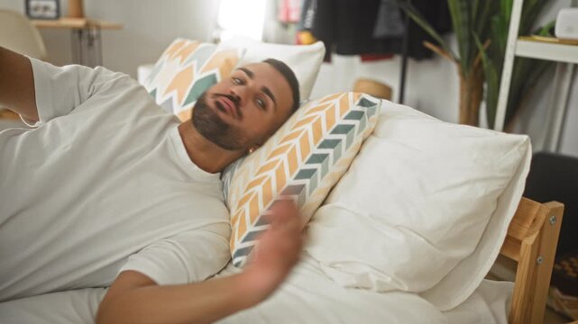 Man lying on bed hugging patterned pillow and turning head on white sheets inside a building; melancholy solitude.