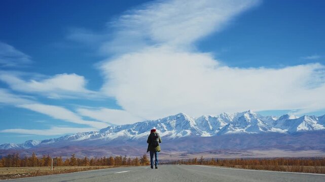 Lone traveler walks a long highway. Mountains stretch behind them, snow-capped and stark. Sky is vast, with wispy clouds drifting. The path leads through open plains under bright sun