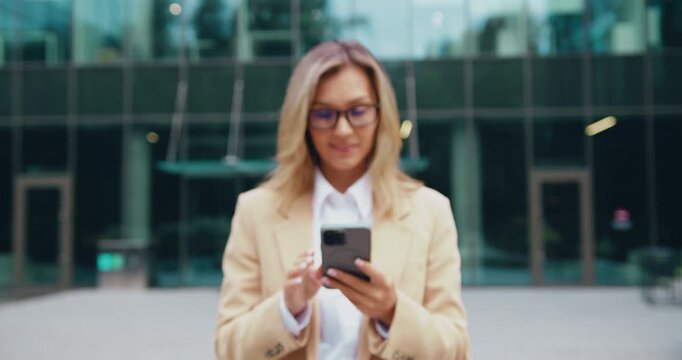 Professional woman standing in front of modern glass office building smiling while using smartphone handheld interaction under soft daylight view from chest up to full body