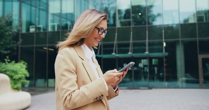 Businesswoman walking outside modern office building checking smartphone with smile and coat moving in breeze, daytime urban plaza with glass facade and greenery, side profile view, professional techl