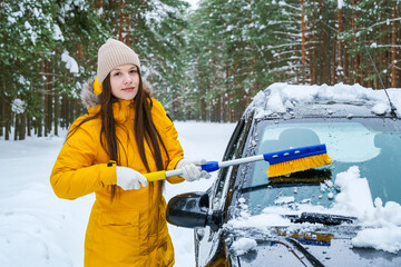 Young woman clearing snow from car with brush in winter forest. Snow removal, vehicle maintenance and safe driving preparation during cold weather concept. © Dmitrii Labrador