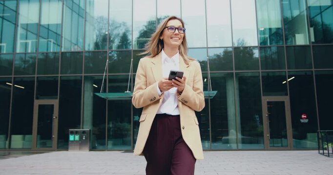 Young professional woman walking toward camera while using smartphone in front of modern glass office building during daylight hours full-length confident stride and smiling expression observed from