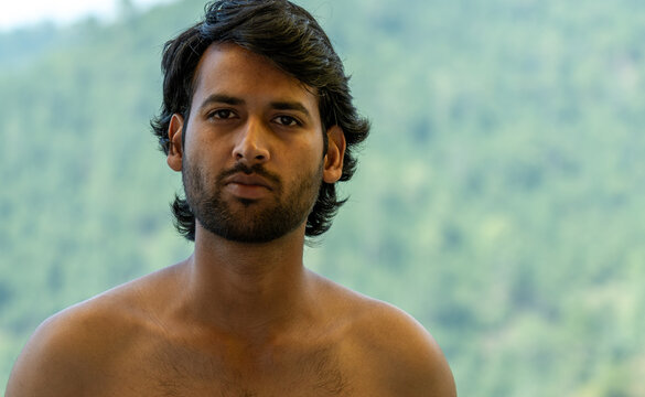 A closeup portrait of a handsome young Indian bearded shirtless man with a serious, confident, and neutral expression looking directly at the camera with an unflinching gaze.