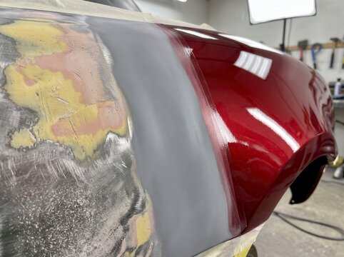 Stages of car body restoration on a vehicle fender in an auto repair shop, showing bare sanded metal, body filler, grey primer, and glossy metallic red finish.