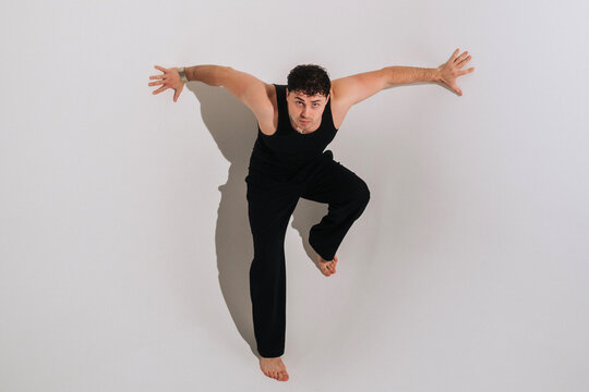 Young man in black tank top balancing on one foot with arms spread, leaning forward against plain wall, barefoot studio portrait with dramatic shadow in