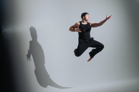 Male dancer midair leap reaching forward with dramatic shadow on studio wall, contemporary performance gesture captured in soft high-contrast light, bare feet