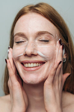 Young woman smiling while massaging cleanser foam into face with eyes closed, close-up portrait expressing relaxation and skincare routine light neutral
