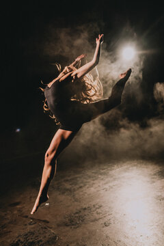 Contemporary dancer midair leap on smoky dim stage under strong backlight, hair and limbs extended in dynamic silhouette of motion and strength and artistic