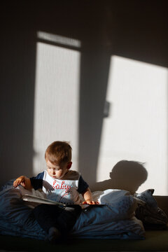 Toddler reading picture book on bed under long window shadows with warm afternoon light,indoor scene with soft contrast and playful silhouette,cozy nursery