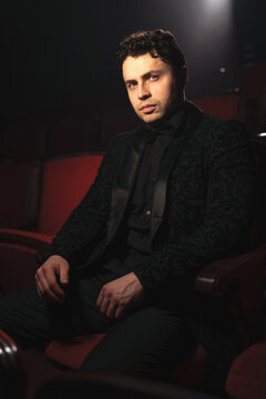 Contemplative young man in ornate black tuxedo jacket and bow tie seated under dramatic theater spotlight, hands resting on knees, moody low key portrait
