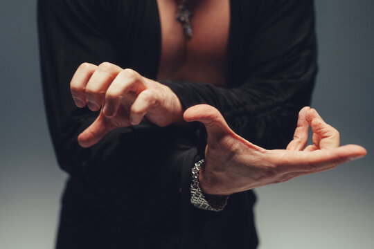 Man adjusting cuff with bracelet and open collared shirt in studio close-up view of hands and chest pendant made visible by soft directional lighting and