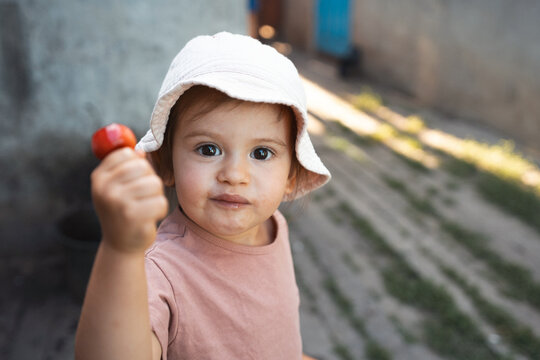 Toddler in sunhat offering small red fruit toward camera with curious expression outdoors, textured stone path background, soft natural light highlighting face