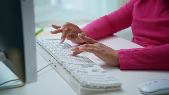 Woman's hands type on white keyboard. Pink sleeves frame focused fingers. Soft light fills clean desk surface. Work, study, or creative task. Professional or casual setting