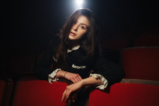 Young woman leaning over red theater seats under single overhead spotlight with contemplative expression and dramatic low light atmosphere that highlights long