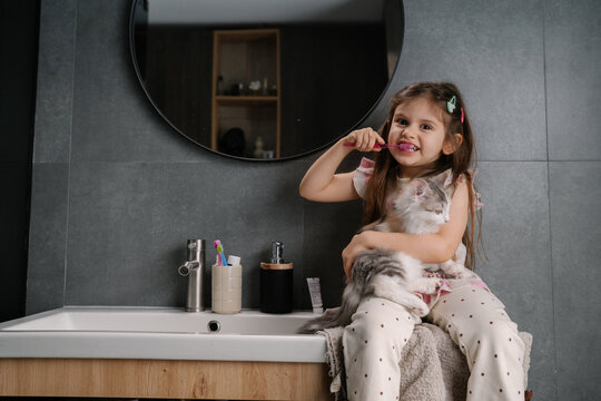 Young girl sitting on bathroom counter holding cat while brushing teeth with pink toothbrush in modern bathroom mirror view, relaxed morning routine with pet