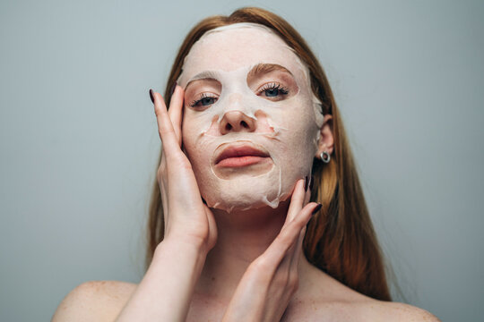 Young woman applying white sheet face mask with hands in minimalist studio portrait, calm self-care moment and skincare routine for beauty and wellness care