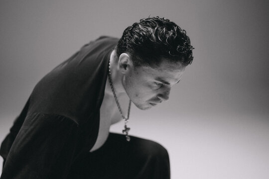 Studio portrait of man leaning forward with focused gaze and pendant necklace, moody monochrome lighting, short curly hair, open dark shirt, dramatic shadowing