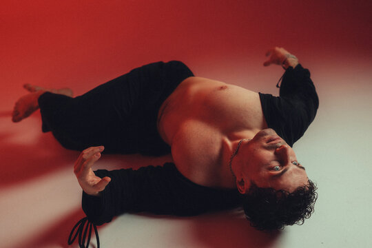 Young man reclining on studio floor with open black shirt exposed chest red dramatic lighting moody fashion portrait