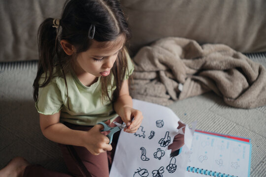 Child cutting paper with scissors while working on craft sheet on couch in cozy living room, focused expression and natural light, hands-on creative activity