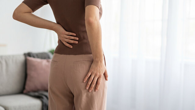 Rear View of Woman Holding Lower Back and Hip Showing Sciatica or Back Pain in Bright Living Room with Copy Space