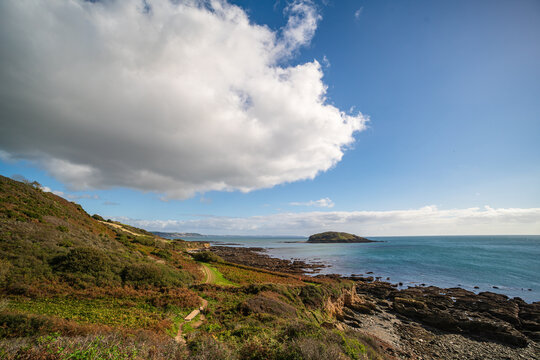 View of the South West Coast Path overlooking the rocky shoreline and Looe Island under a bright blue sky with large clouds in Looe, England, United Kingdom.