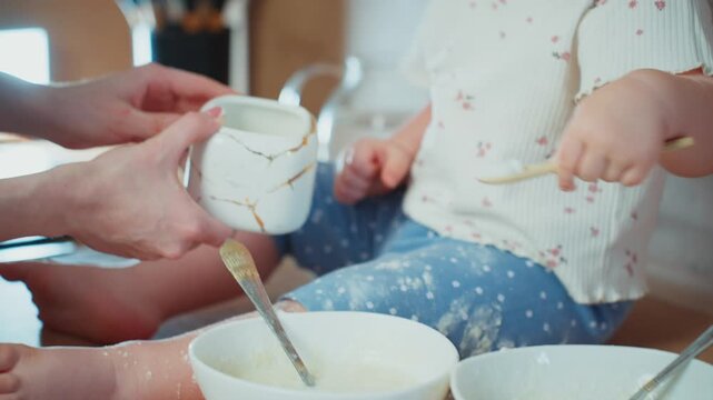 Soft light bathes a cozy kitchen scene. Child's small hands grip a chipped mug. Adult's hands gently guide the child's cup. Bowl of flour sits beside another bowl