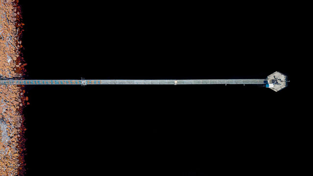Aerial view of a long narrow pier extending from a rocky shoreline into the dark water of a dam, ending in a hexagonal platform in Grabouw, Western Cape, South Africa.