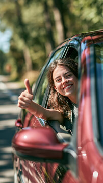 a young woman in a red car. She is smiling and giving a thumbsup with her right hand. Her long, wavy brown hair falls over her shoulders. She wears a gray shirt.