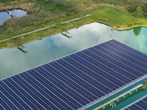 Aerial view of a floating solar farm installed on a calm pond, with rows of photovoltaic panels reflecting the sky, surrounded by green grassy banks and trees in Jurmala, Latvia.